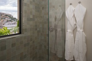 Una cabina doccia in vetro con vista sulla città, una finestra e due accappatoi bianchi appesi al muro.
