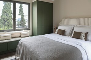Una camera da letto con un letto grande, una panca finestrata con cuscini, un'armadio verde e vista sugli alberi.