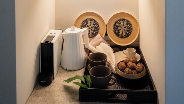 Quarto com planta em vaso, bandeja com canecas e biscoitos, cafeteira e um alto-falante.