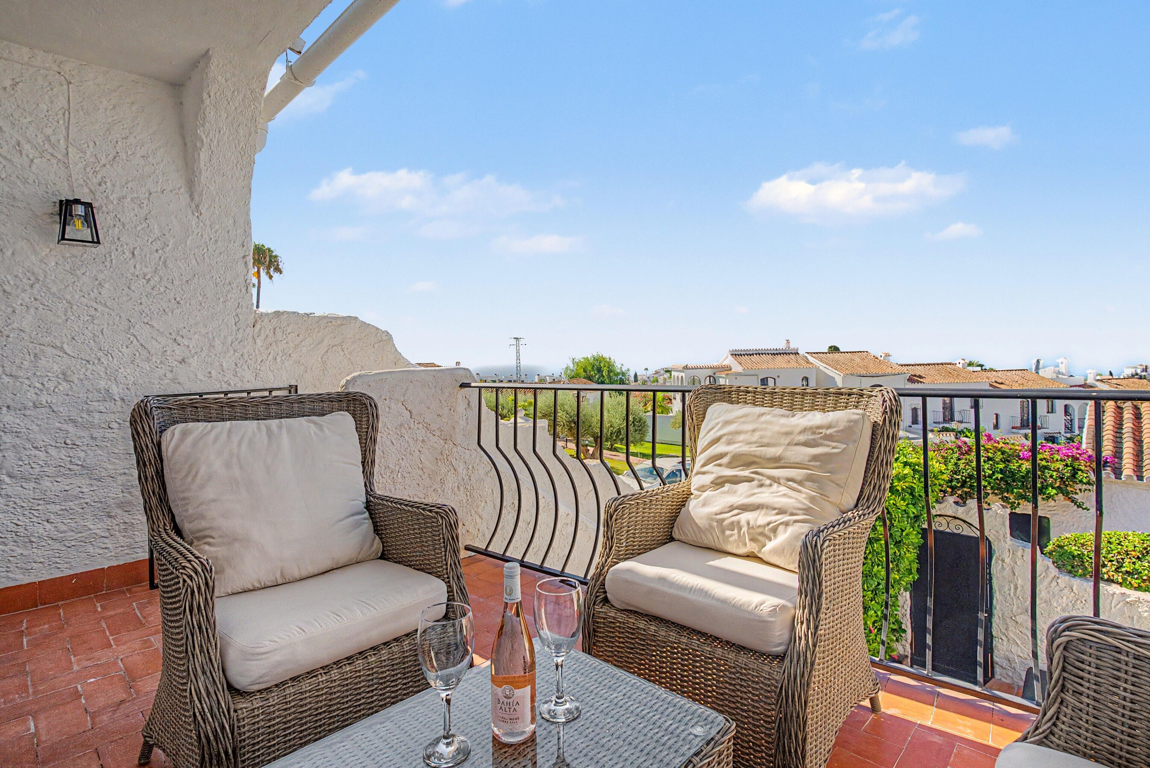 A balcony with wicker furniture, a table with glasses and a bottle, and a view of buildings and greenery.
