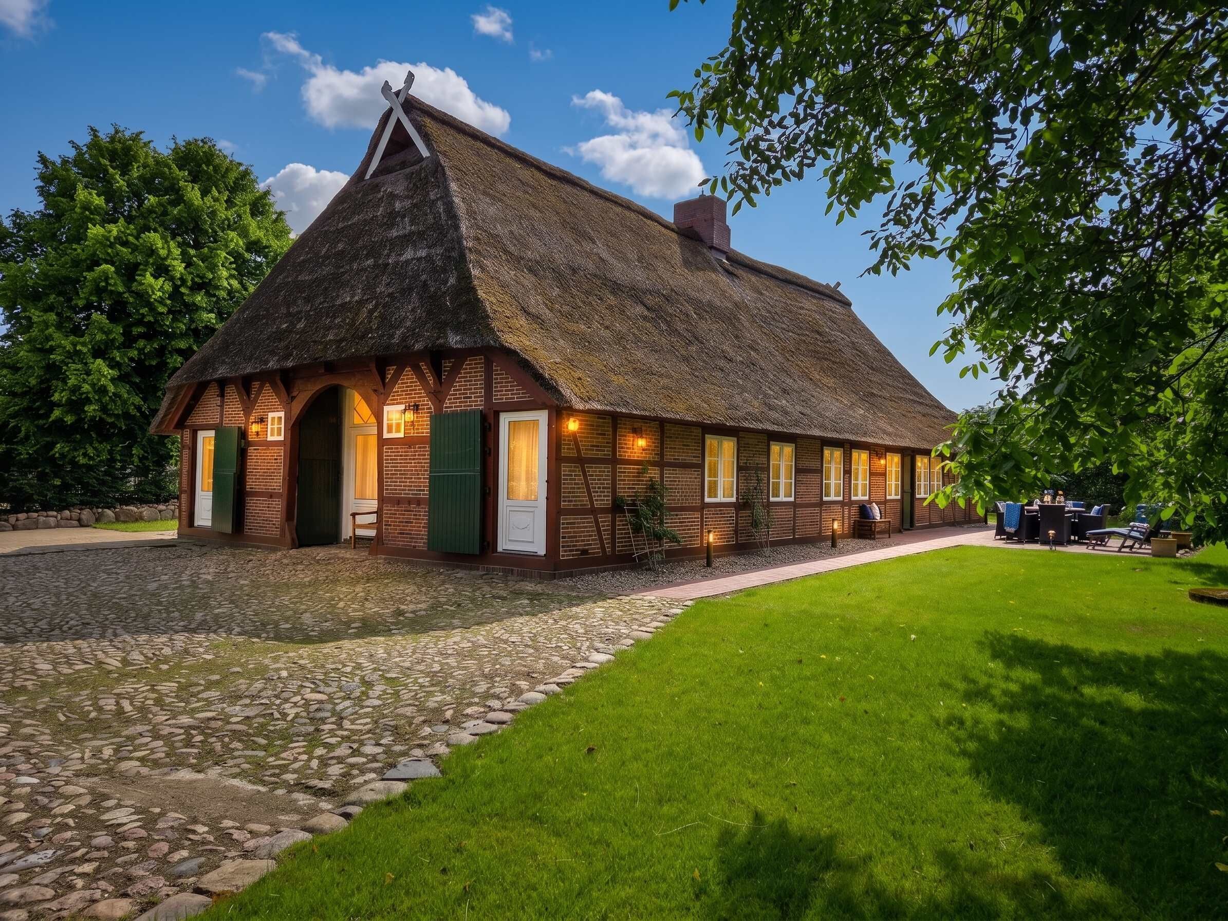 Une maison de campagne traditionnelle à toit de chaume, avec un chemin de pierre menant à l’entrée, entourée d’une pelouse bien entretenue et d’arbres.