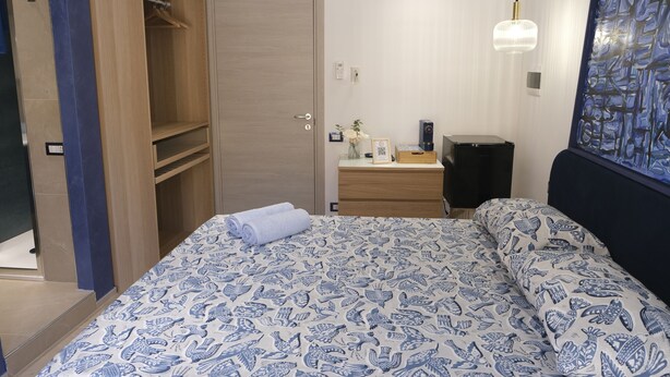 A bedroom with a bed, a wardrobe, a bedside table with a lamp, and a wall with a patterned artwork. - Room with all the amenities (Termini Imerese)