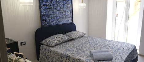 A bedroom with a bed, a painting, a nightstand, a lamp, and an air conditioning unit.