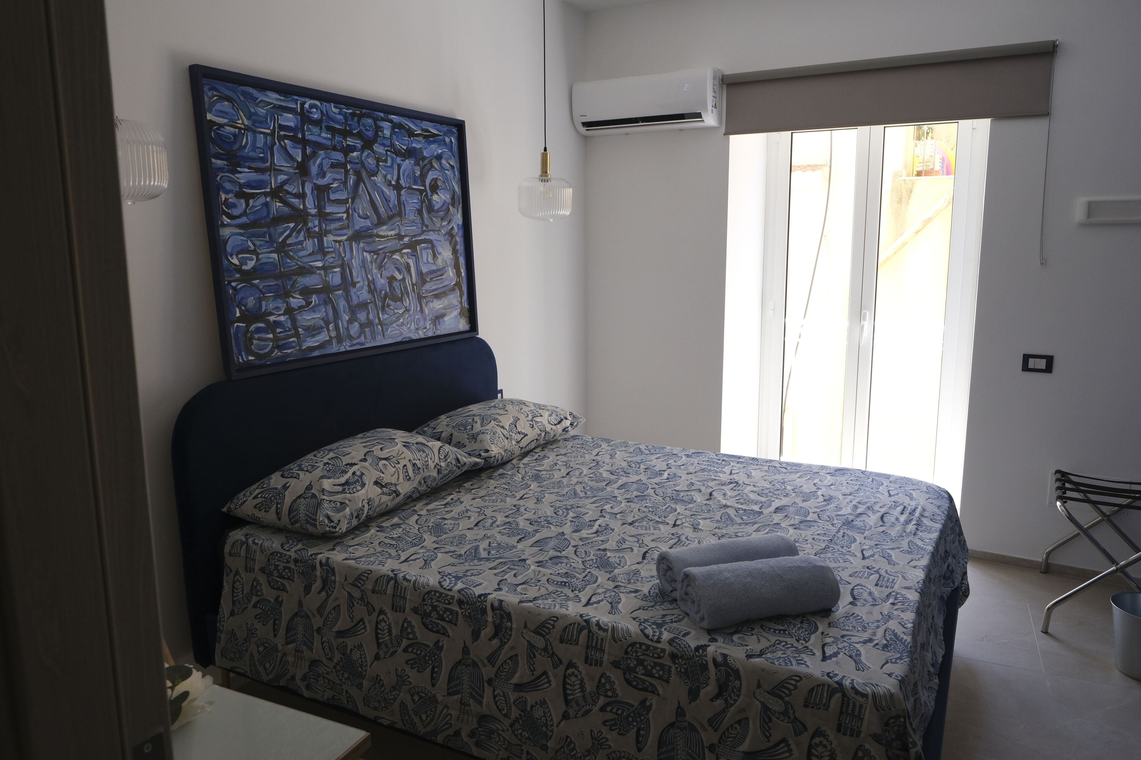 A bedroom with a bed, a painting, an air conditioning unit, and a window with curtains.