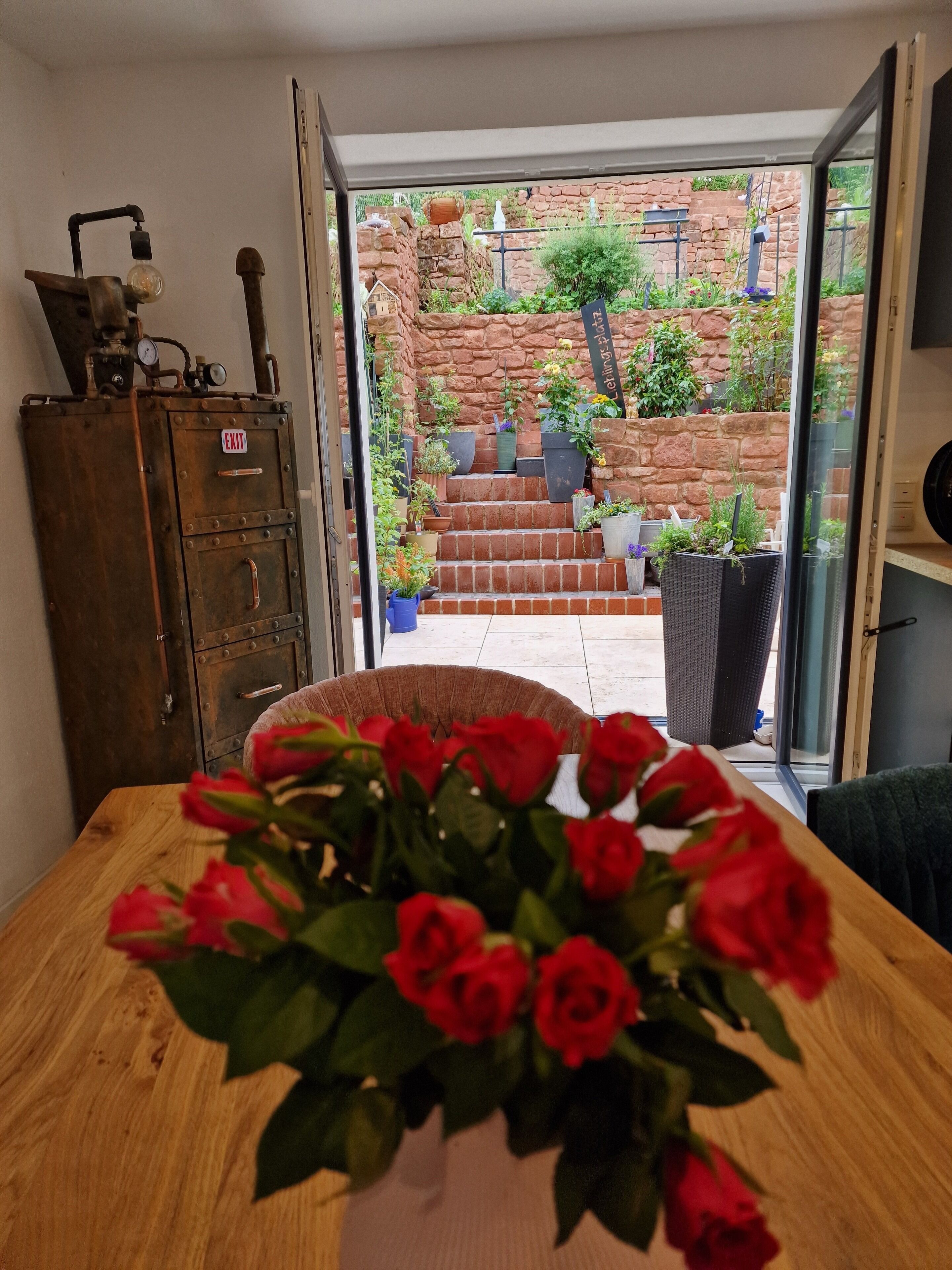A room with a wooden table, a vase of red roses, a vintage metal cabinet, and a view of a stone wall and plants through an open door.