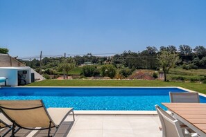 A rectangular swimming pool with clear blue water, surrounded by a tiled patio with two lounge chairs and a wooden table. - Mosteiros Escape with Pool & BBQ by Lisbeyond (Mosteiros)