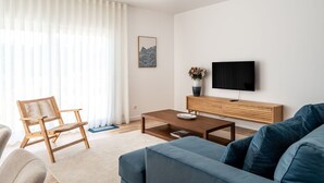 Een moderne woonkamer met een bank, een houten salontafel, een tv op een houten standaard, een rieten fauteuil en een groot raam met dunne gordijnen.