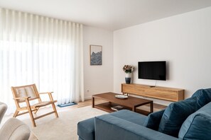 A modern living room with a sofa, a wooden coffee table, a TV on a wooden stand, a wicker chair, and a large window with sheer curtains. - Mosteiros Escape with Pool & BBQ by Lisbeyond (Mosteiros)