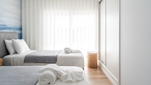 Habitació d'hotel moderna amb dos llits, un gran finestral amb cortines translúcides, una làmpada de nit i una taula rodona petita.