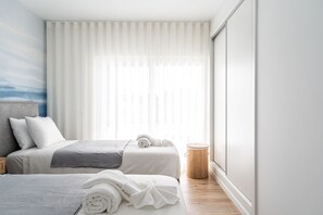 Ein modernes Hotelzimmer mit zwei Betten, einem großen Fenster mit transparenten Vorhängen, einer Nachttischlampe und einem kleinen runden Tisch.