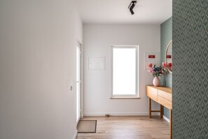 Ein Flur mit Holzboden, einer Tür, einem Fenster, einem an der Wand befestigten Schild und einer Holzbänkl mit einer Blumenvase.
