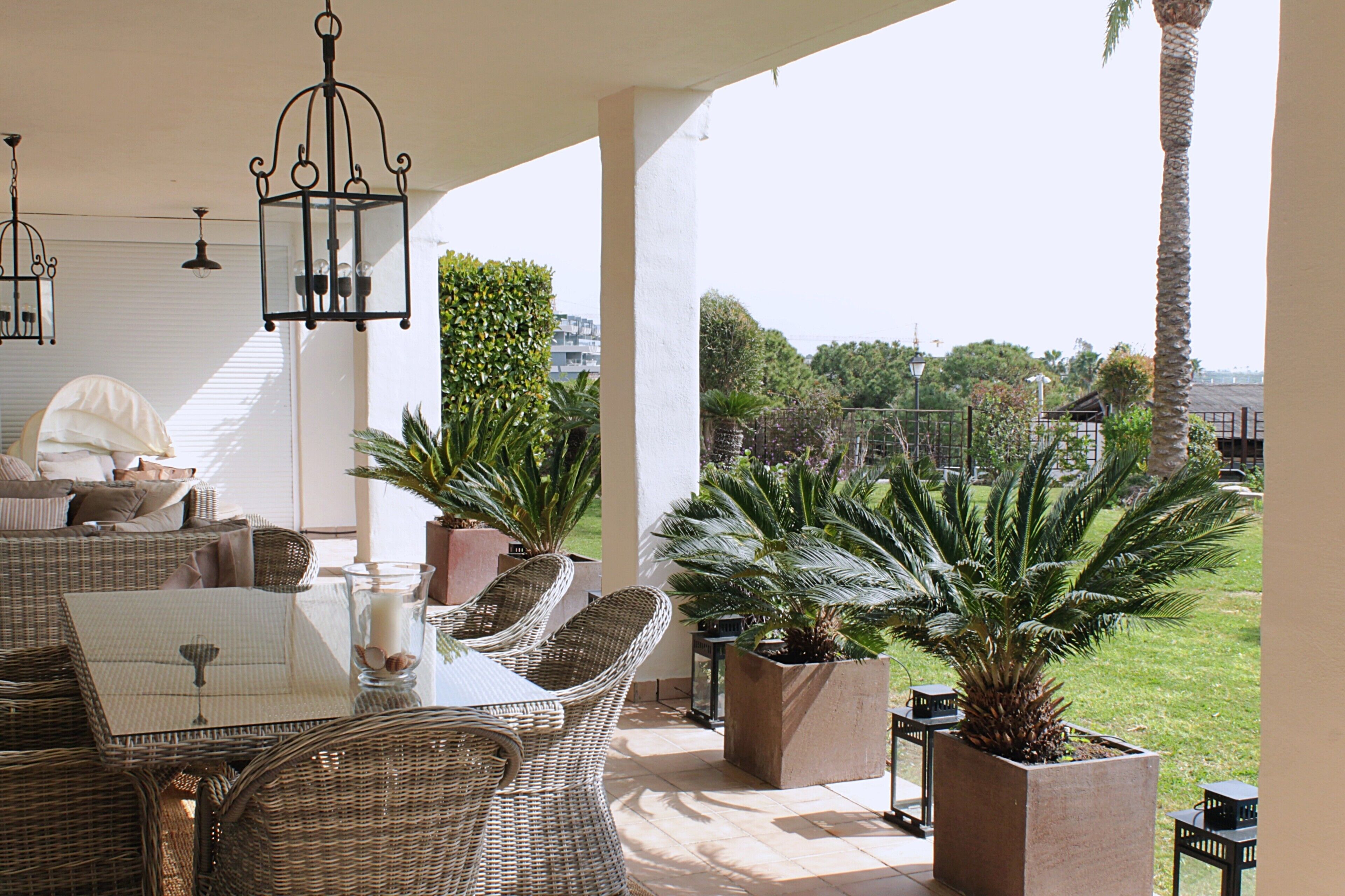 A covered outdoor seating area with wicker furniture, potted plants, and hanging lanterns.