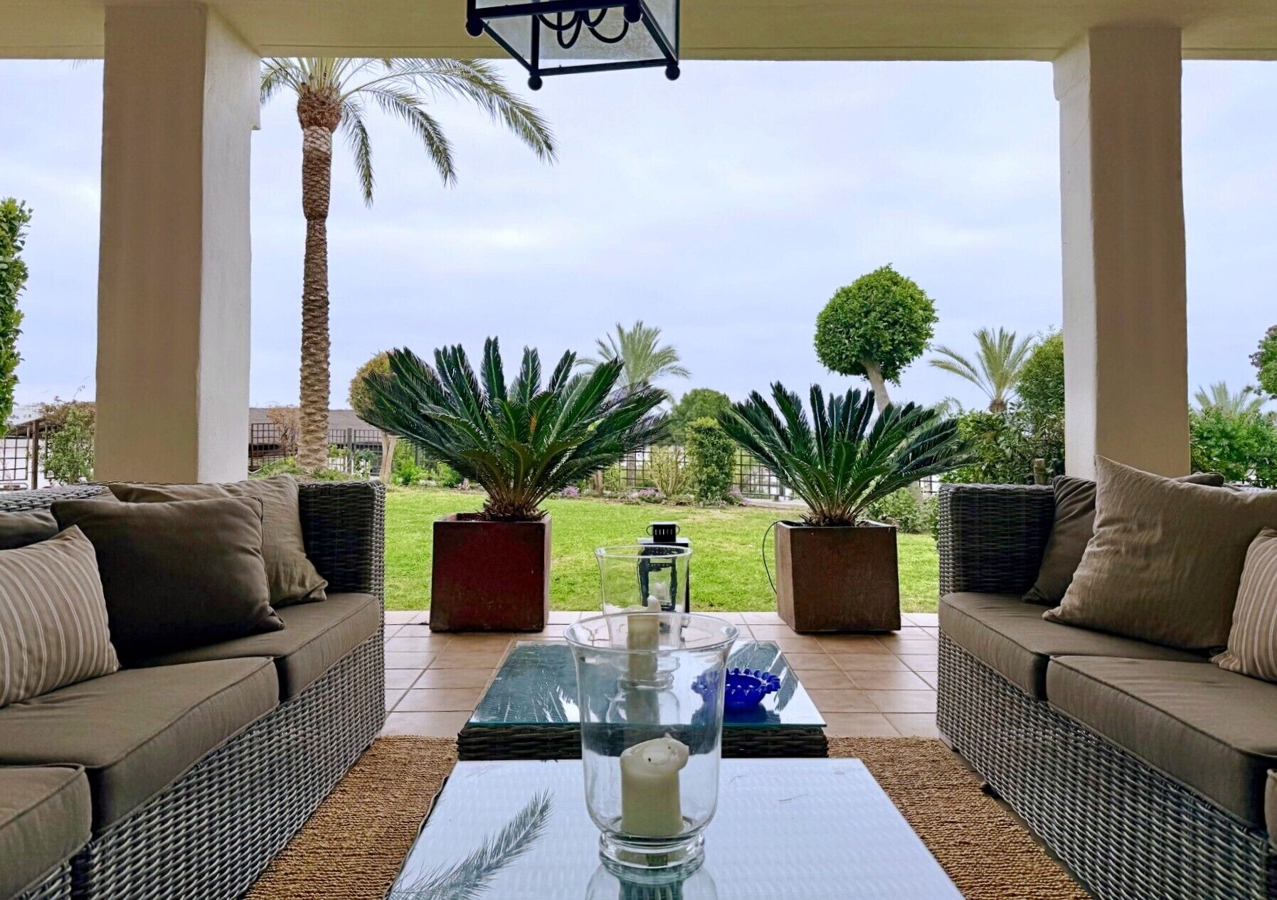 Een overdekt terras met twee banken, een glazen tafel en potplanten.