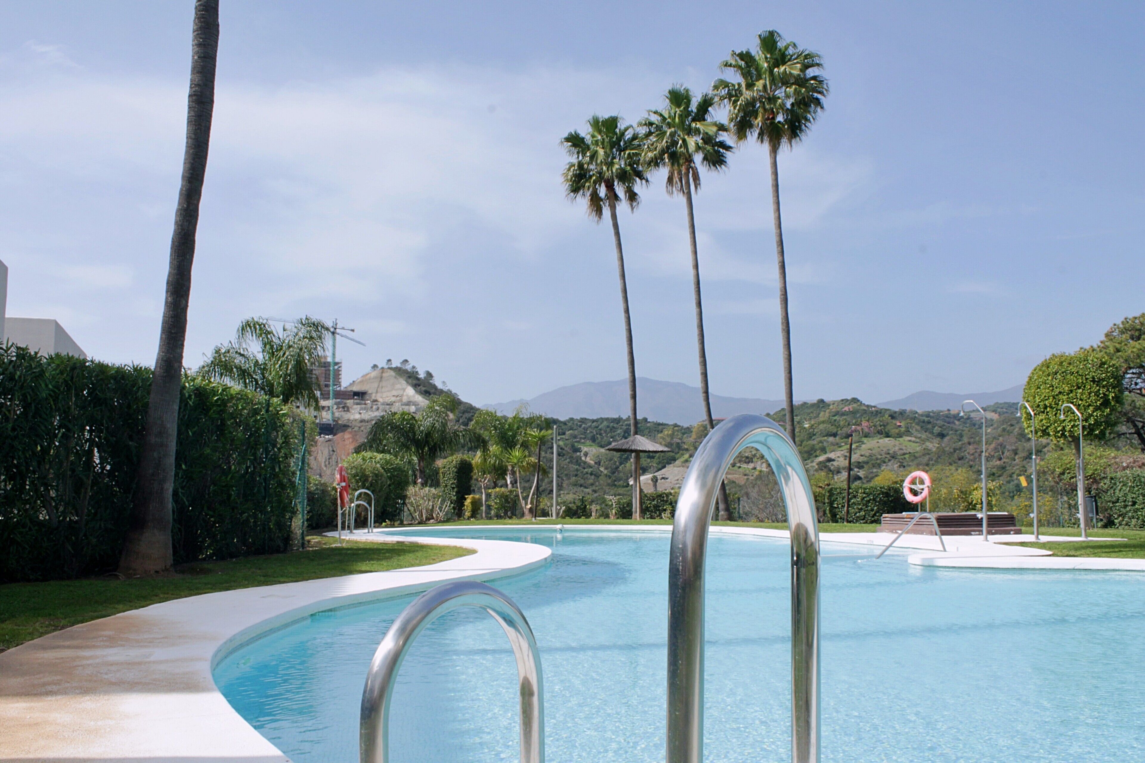 A swimming pool with a curved edge, surrounded by palm trees and a hilly landscape in the background.