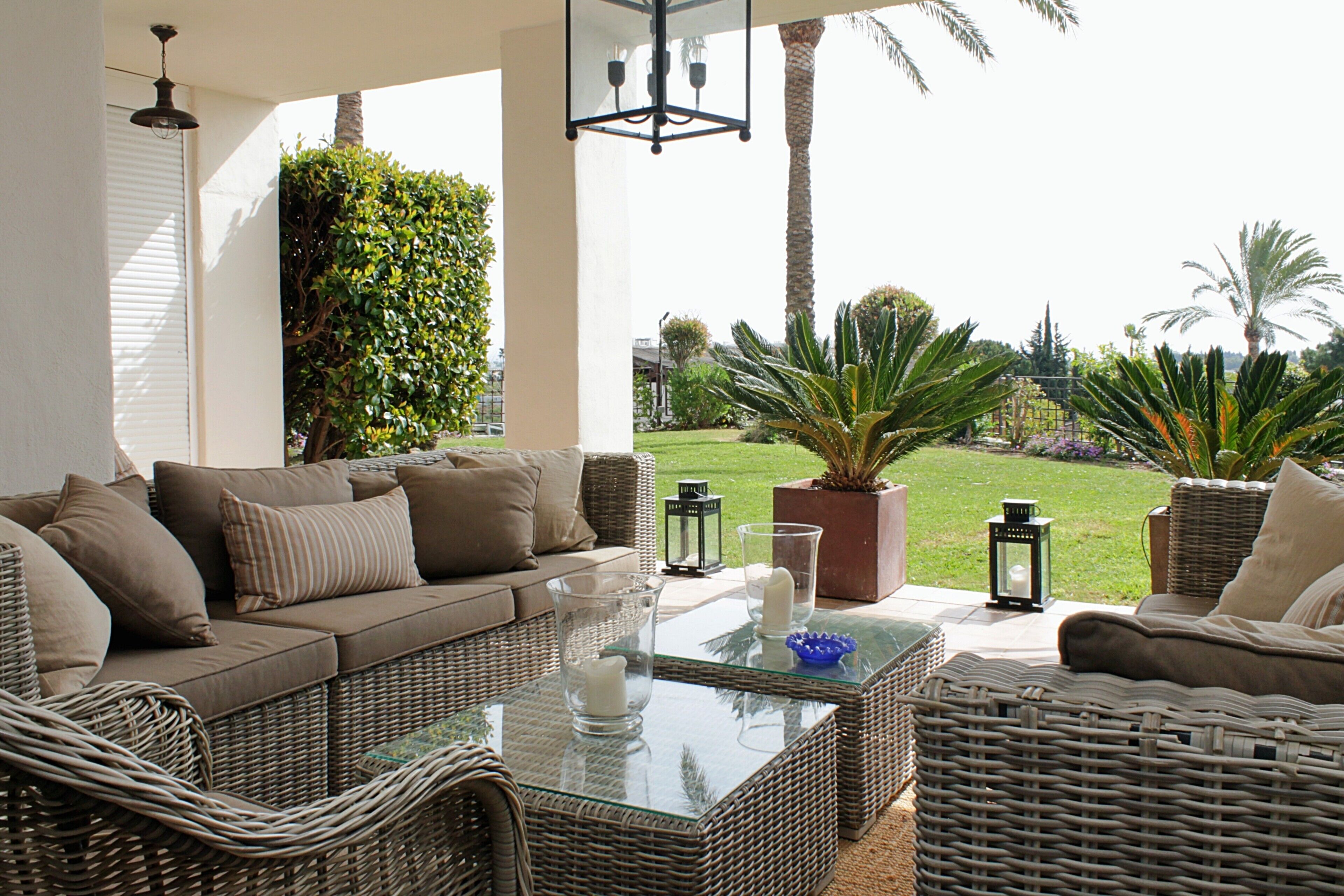 A sunlit outdoor seating area with a wicker sofa, a glass-top coffee table, and potted plants.