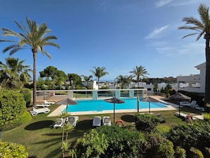 A pool area with lounge chairs and umbrellas, surrounded by palm trees and greenery. - Garden Apartment. Golf by the Sea in Estepona (Estepona)
