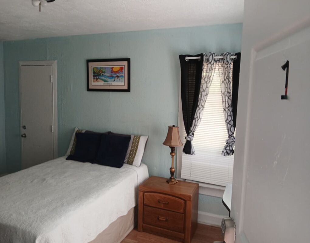 Une chambre à coucher comprenant un lit, une table de chevet, une lampe, une fenêtre avec des rideaux, une porte et un tableau accroché au mur.