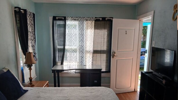 Ein Schlafzimmer mit Bett, Schreibtisch, Fernseher und einer zur Außenwelt führenden Tür.