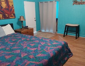 Een slaapkamer met een bed, nachtkastje, lamp, stoel, deur en gordijn.