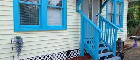 Une maison avec un escalier bleu menant à une porte, une clôture blanche en bois et un petit jardin agrémenté de paillis rouge et de fleurs violettes.