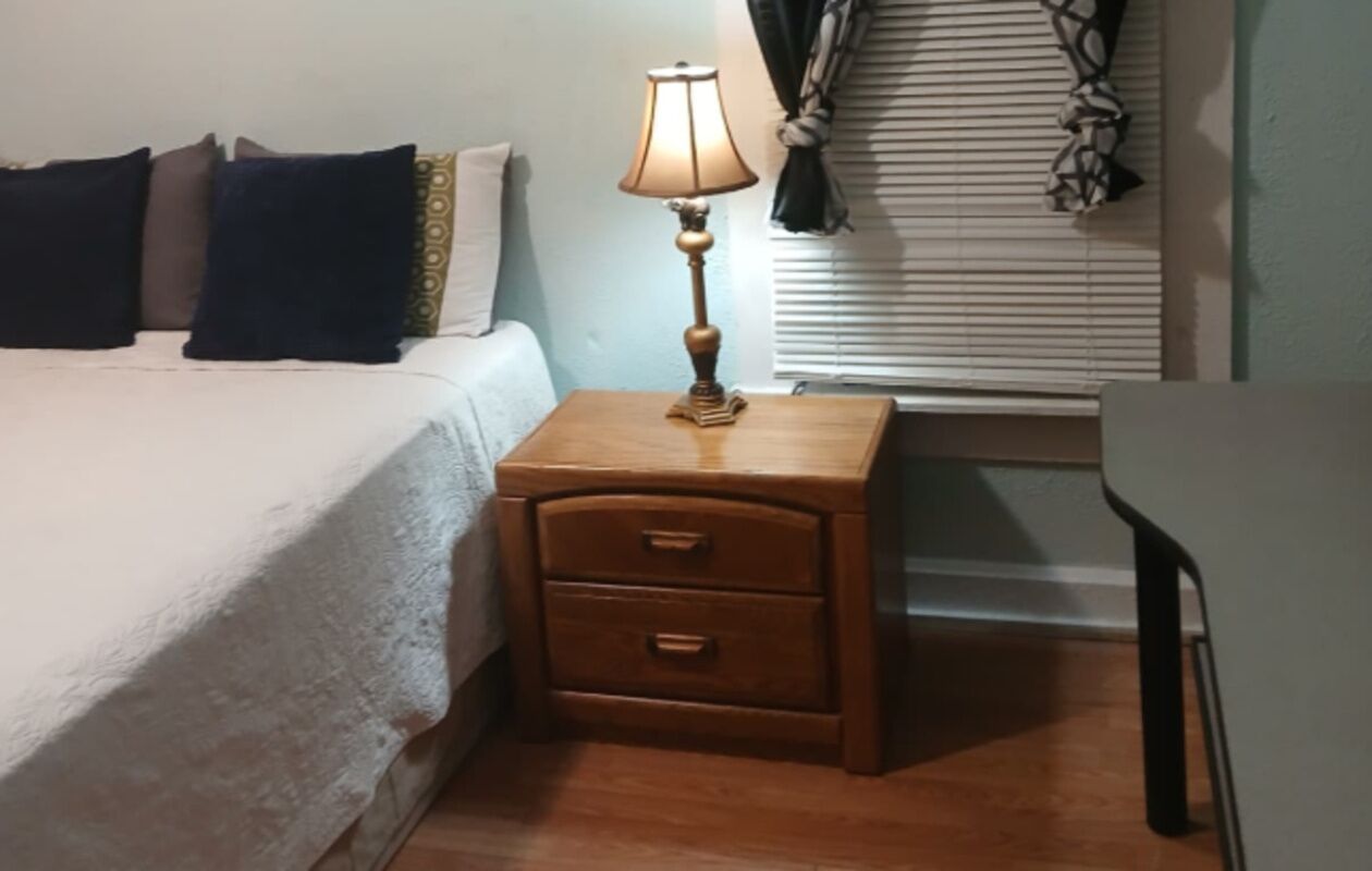 Una cama con edredón blanco, una mesita de noche de madera con lámpara y un cajón pequeño.