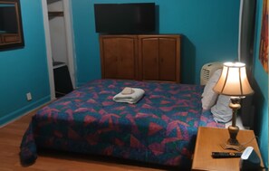 Un dormitorio con una cama, un televisor encima, un armario, una lámpara en una mesita de noche y un control remoto en la mesa.