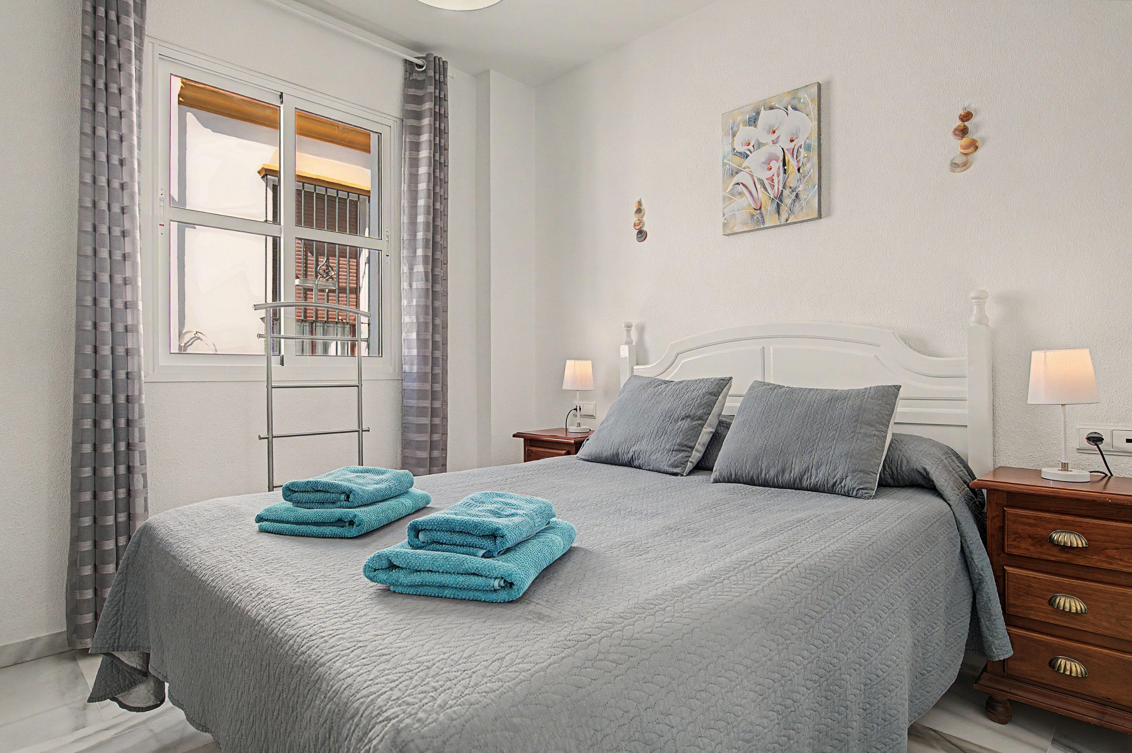 A hotel room with a bed, two bedside tables, a nightstand, a window with curtains, and a wall-mounted artwork.