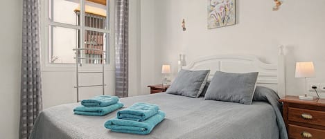 A hotel room with a bed, two bedside tables, a nightstand, a window with curtains, and a wall-mounted artwork.