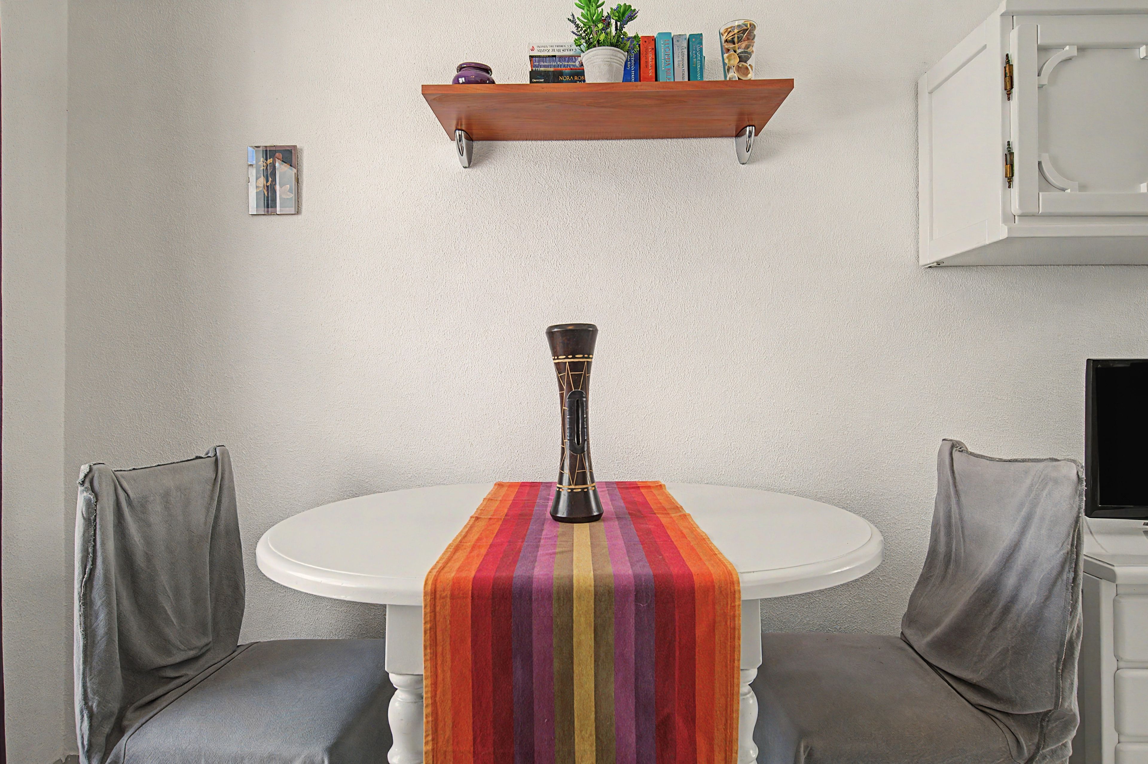 A round dining table with a colorful striped table runner, two grey chairs, a decorative vase, and a shelf with books and decorative items.