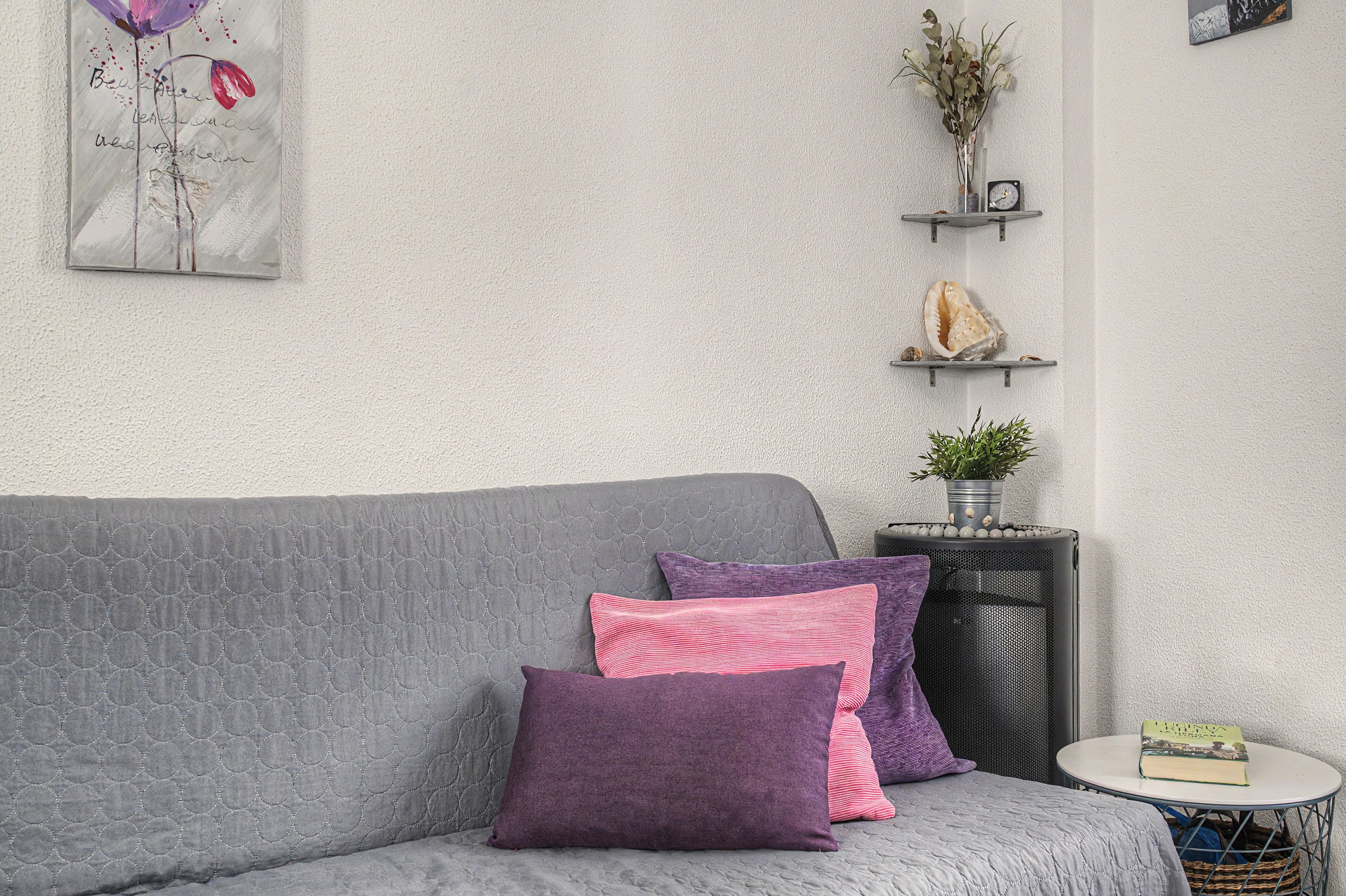 Un divano grigio con cuscini viola e rosa, un tavolino rotondo con un libro e oggetti decorativi su una parete bianca con texture.