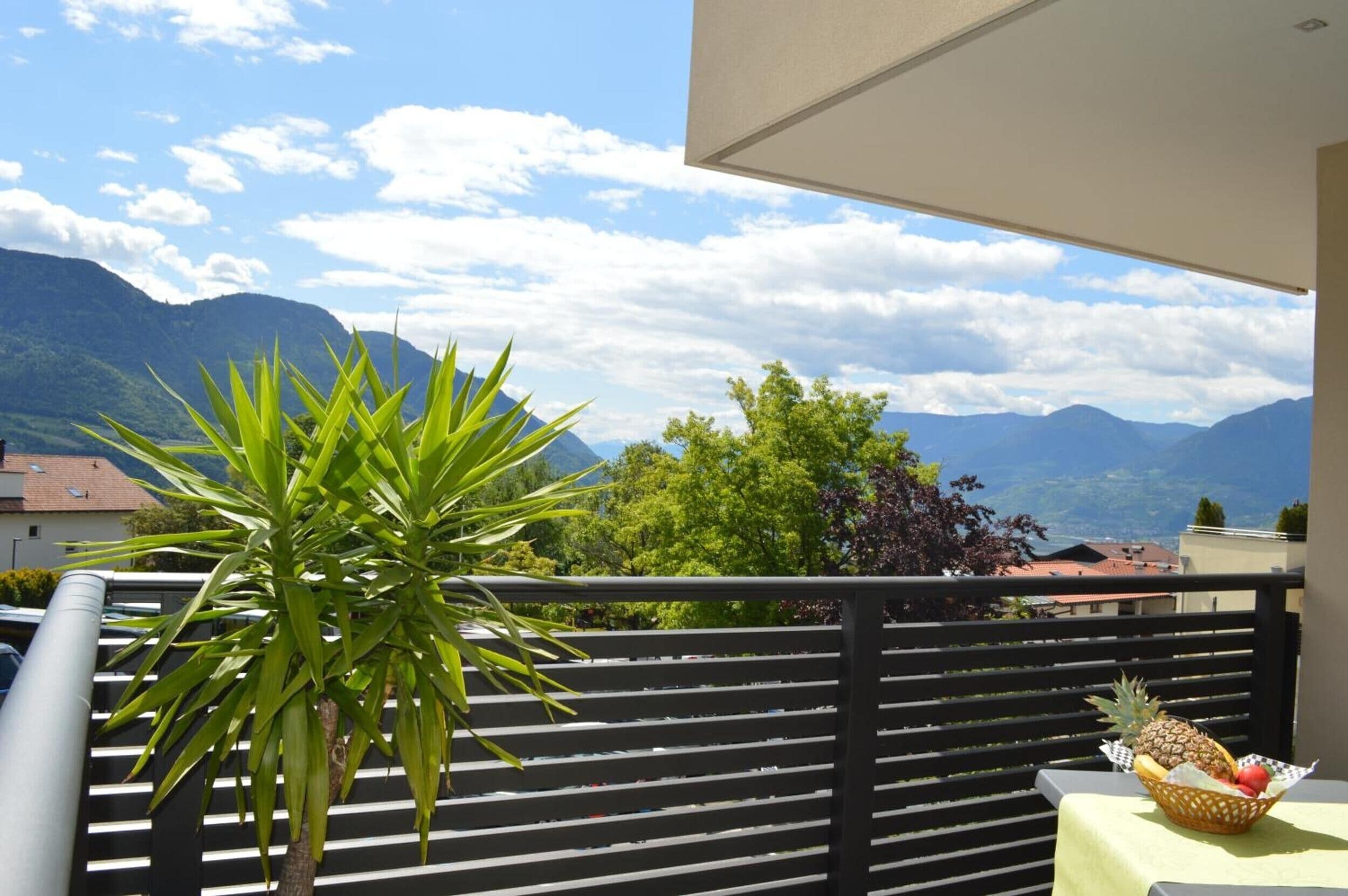 Un balcon avec vue sur les montagnes, une plante en pot et une table sur laquelle se trouve un panier de fruits.