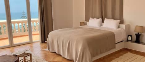 A bedroom with a large bed, a balcony with a view, and a dining area with a table and chairs.