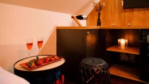 Une pièce chaleureuse avec un meuble en bois, une petite table sur laquelle se trouve un plateau de fraises et deux verres remplis d’une boisson rouge, une bougie et un téléviseur fixé au mur.