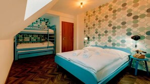 Une chambre avec des lits superposés, un lit simple, un plancher en bois disposé en chevrons, un mur décoré d’un motif et une petite table avec une lampe.