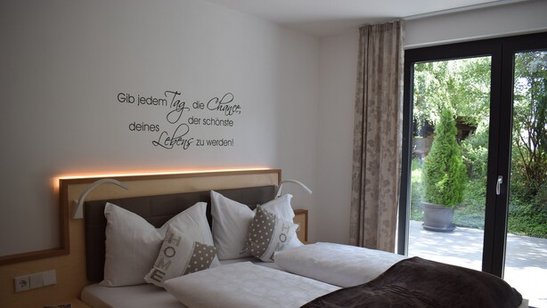 Un dormitorio con cama, almohadas, una mesita de noche, una pared con una cita inspiradora y vistas a la vegetación a través de una ventana.