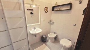 Et badeværelse med toilet, bidet og vask. Der er en lille skraldespand og en hylde med pynteting.
