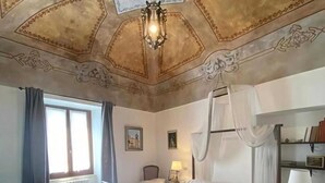 Et soveværelse med himmelseng, skrivebord, stol, et vindue med gardiner og et loft med dekorative mønstre.