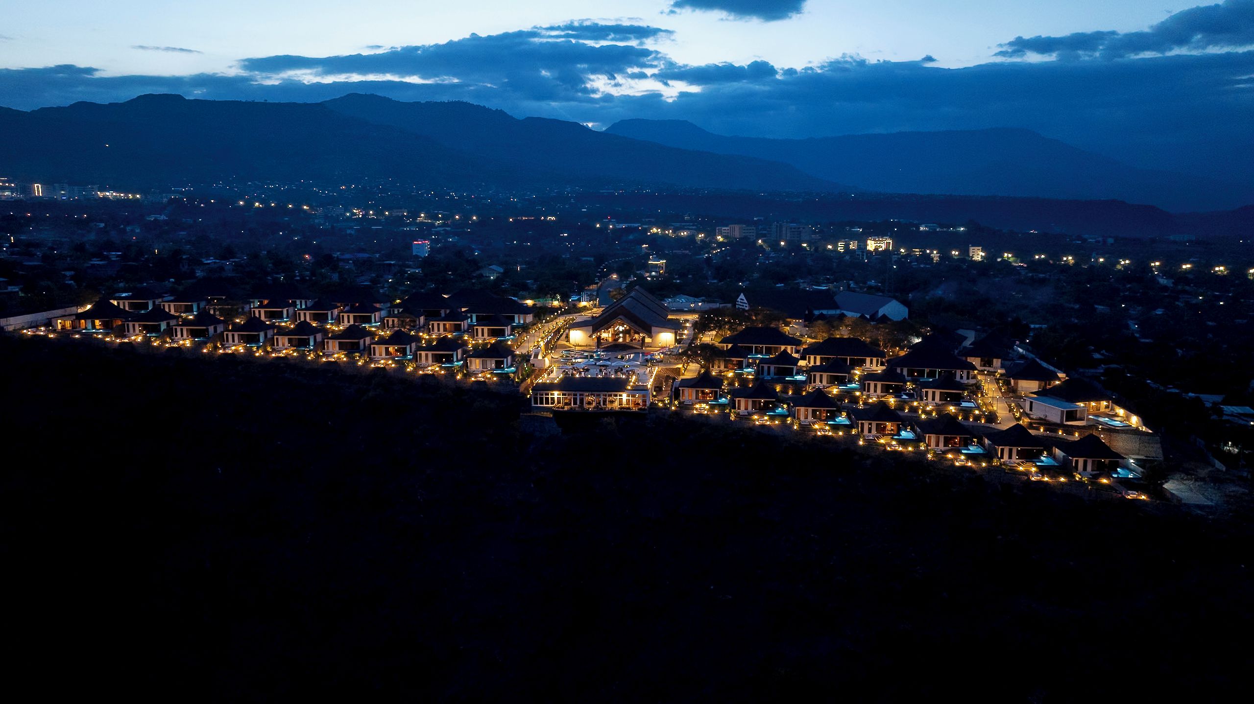 A residential area with houses and buildings at dusk, featuring a hilly landscape in the background.