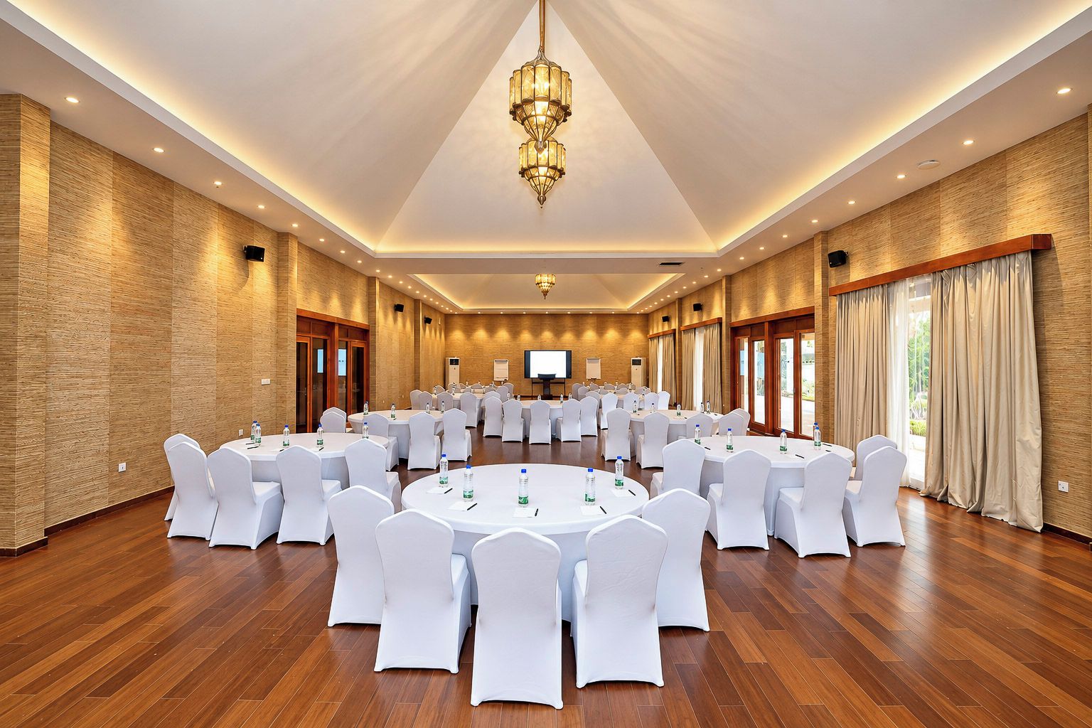 Un salón de conferencias con mesas redondas, sillas blancas y una pantalla grande al frente.