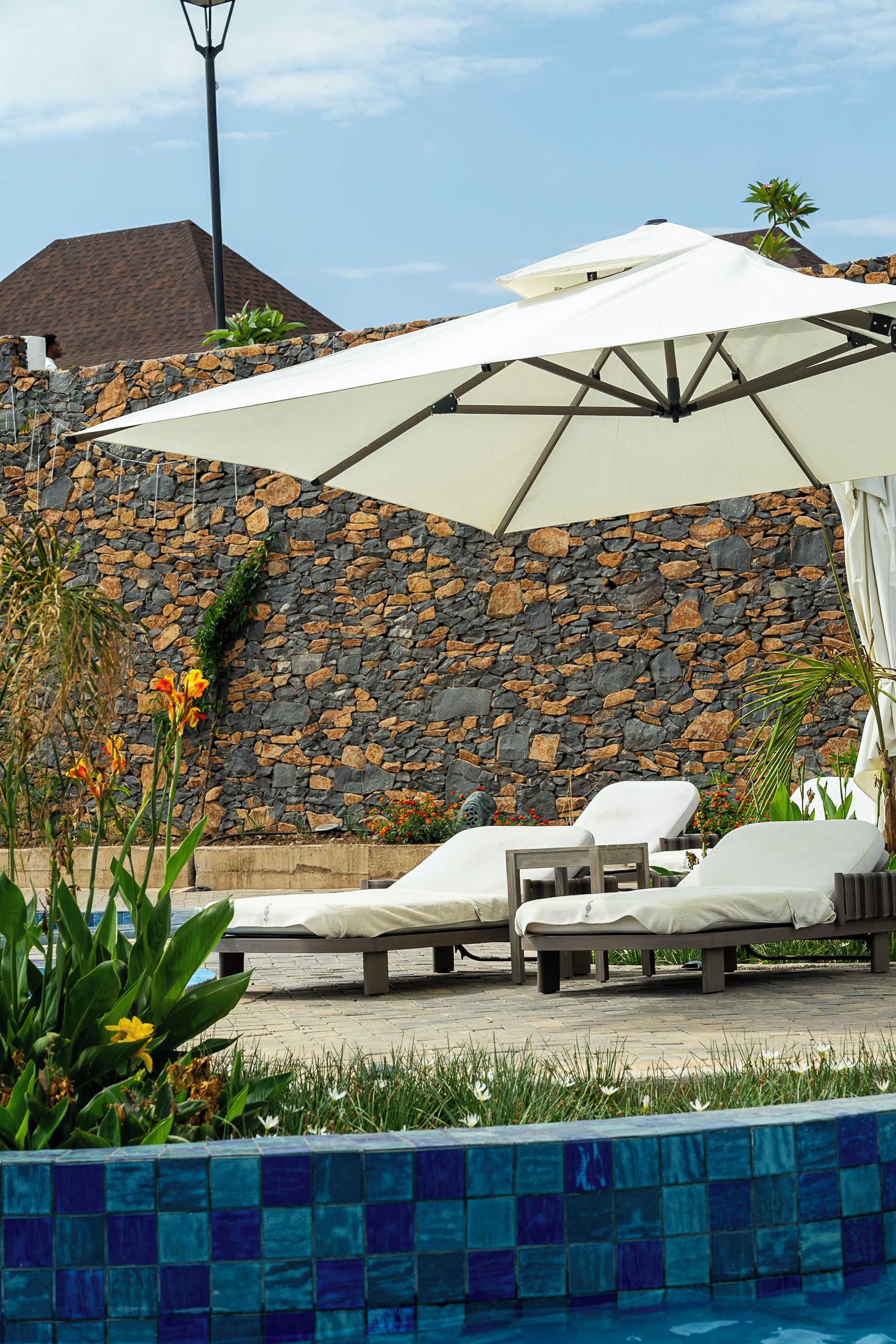 A poolside area with lounge chairs and a large umbrella, set against a stone wall with a tiled pool edge.