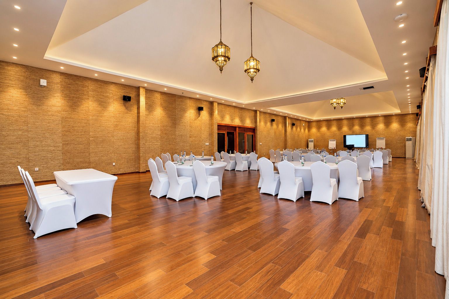 A large conference room with multiple round tables and chairs, wooden flooring, and modern lighting fixtures.