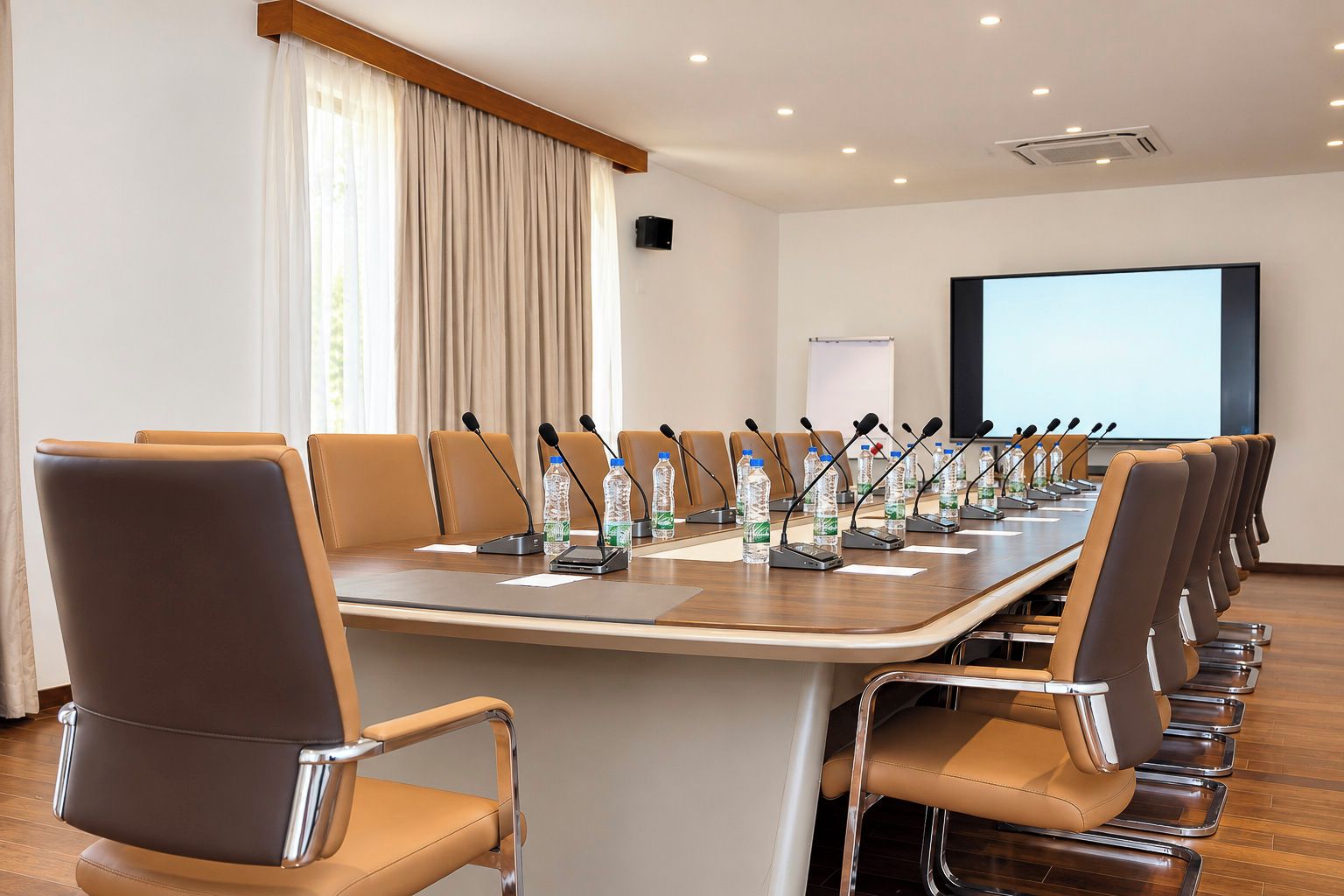Una sala de conferencias con una mesa larga, varias sillas, micrófonos y botellas de agua.