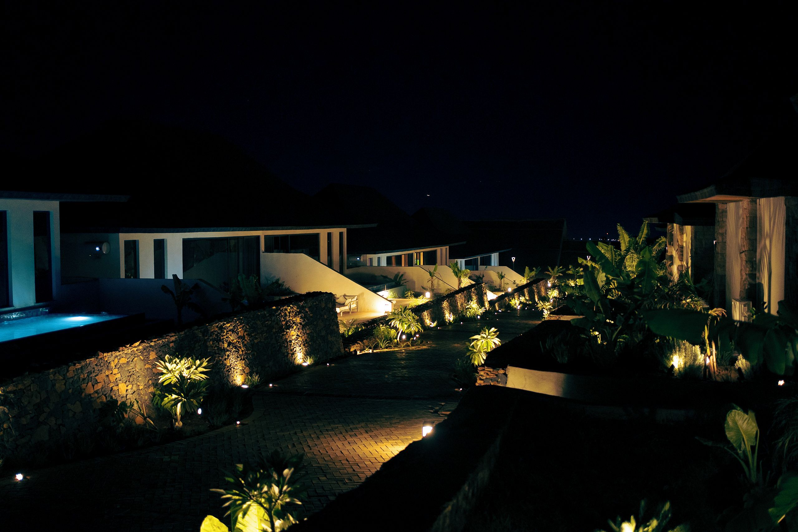 Vista nocturna de un complejo turístico con piscina, muros de piedra y plantas tropicales.