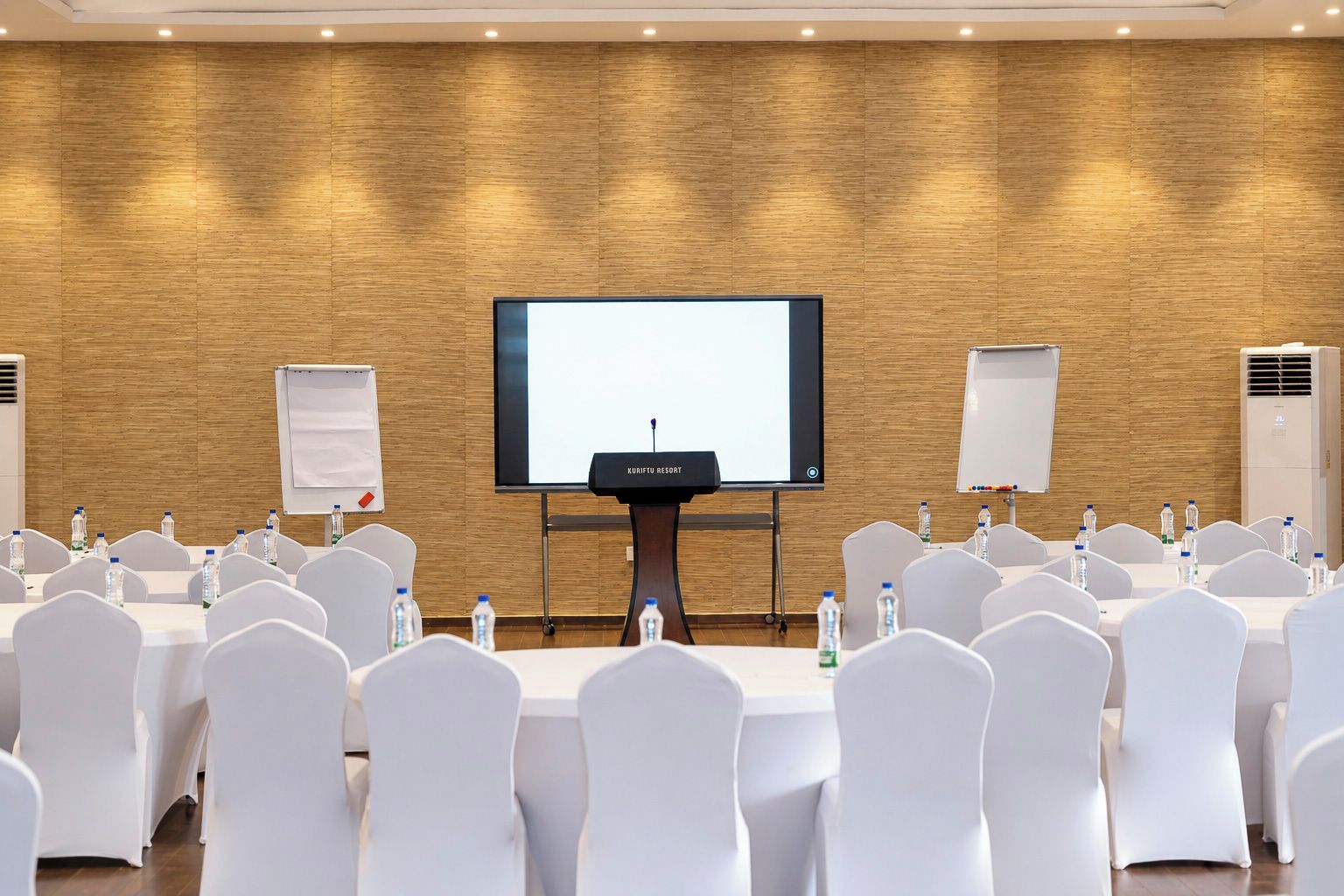 A conference room with a large screen, white chairs, tables, and water bottles.