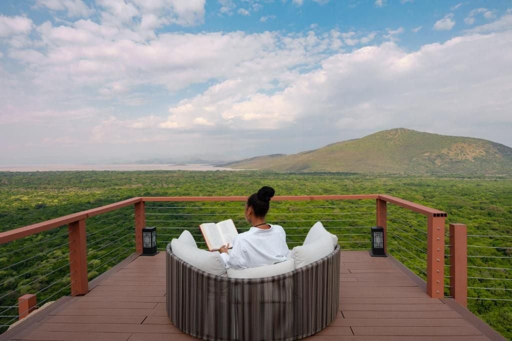 Una persona sentada en un banco circular, leyendo un libro, con vistas a un paisaje pintoresco con colinas y una masa de agua.