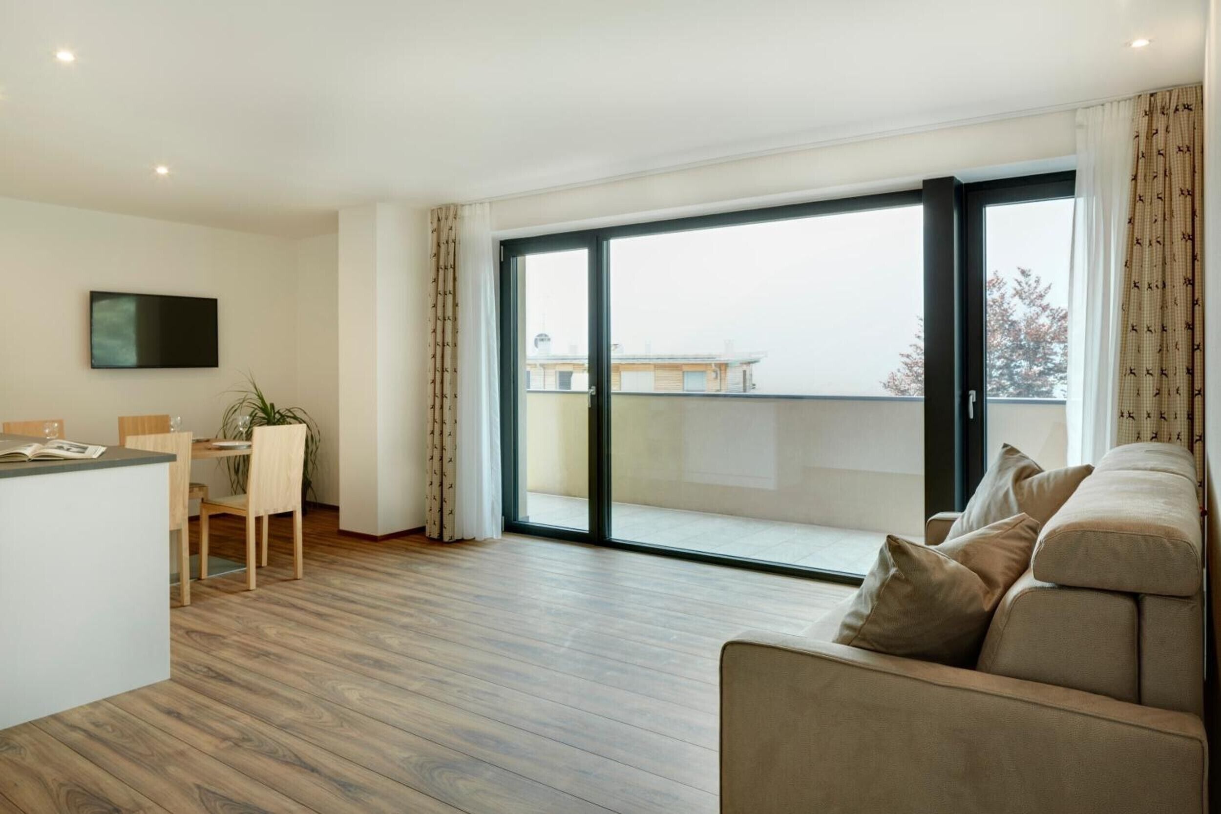 A modern living room with a sofa, a dining area with chairs, a TV mounted on the wall, and a large sliding glass door leading to a balcony.