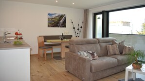 A modern living room with a sofa, a dining area with a table and chairs, a kitchen with a sink and counter, and a large window with a view. - Apartment 'Residence Ladurn Herz' with Mountain View, Balcony and Wi-Fi (Tirol)