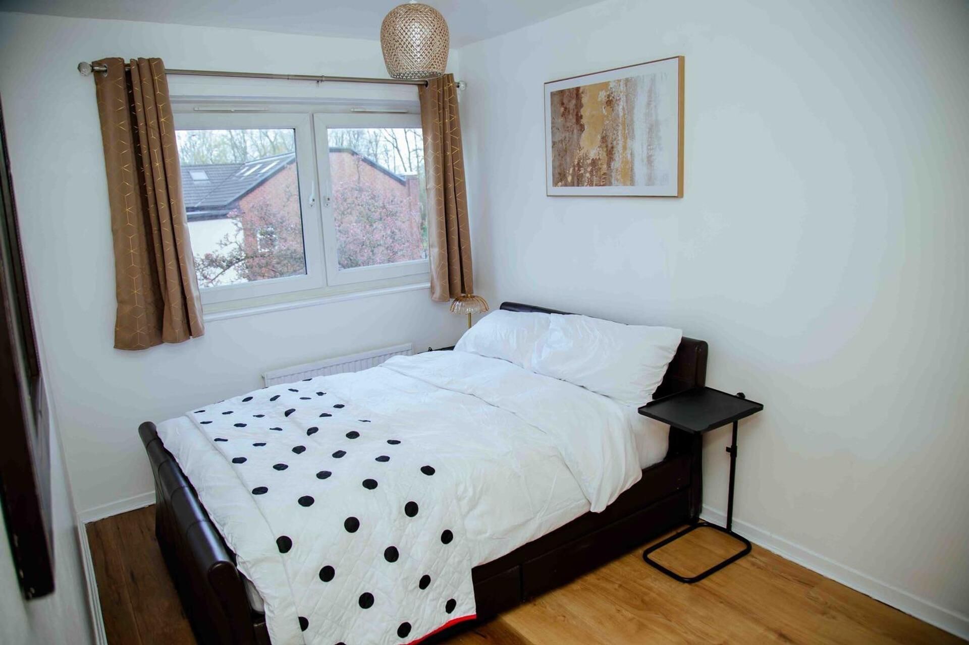 Een slaapkamer met een bed, een nachtkastje, een raam met gordijnen en een kunstwerk aan de muur.