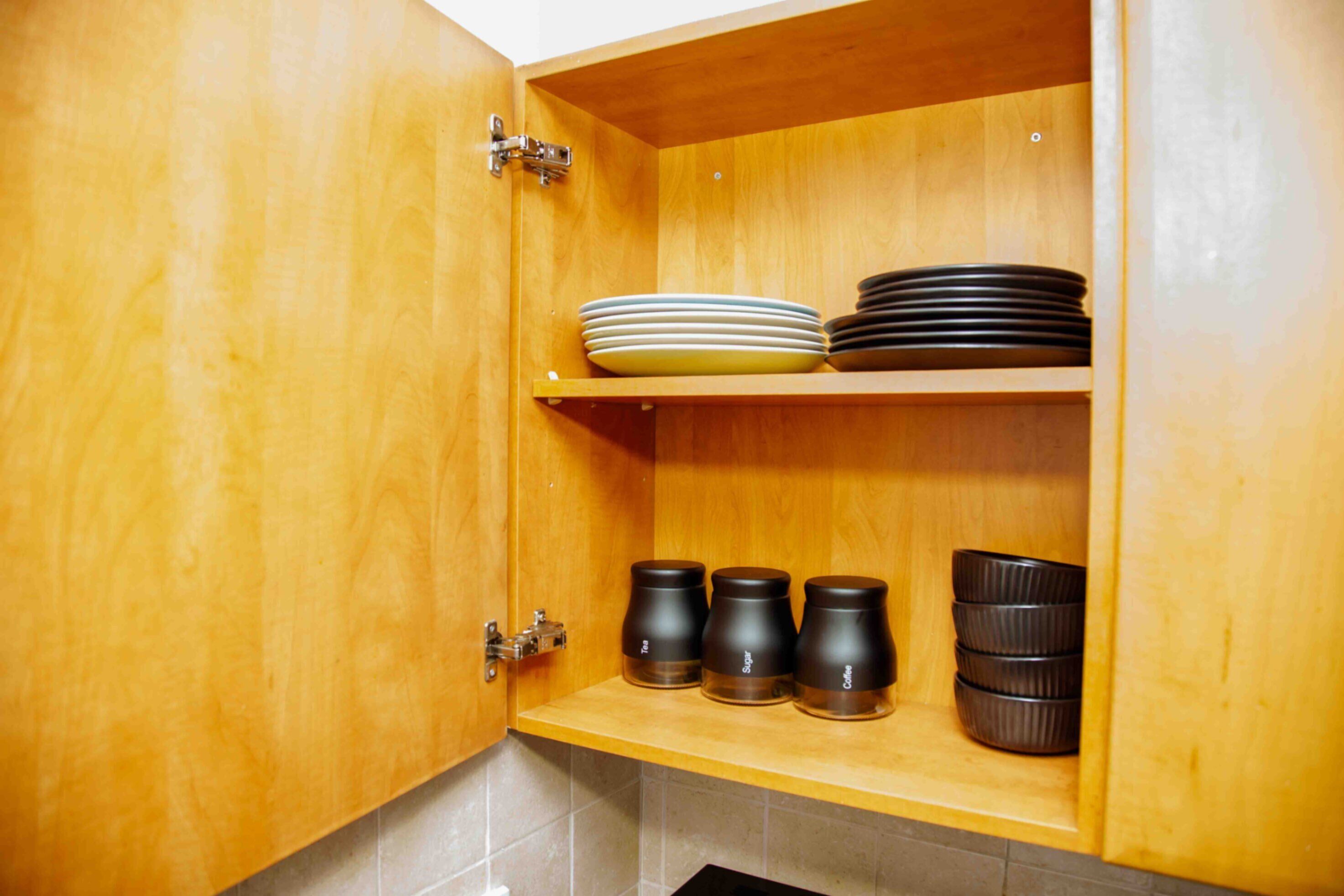 A wooden kitchen cabinet with two shelves; the top shelf holds stacked plates, and the bottom shelf contains black mugs.