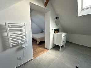 Ein modernes, kompaktes Schlafzimmer mit schrägem Dach, integriertem Heizkörper, Nachttisch mit Spiegel und kleinem Fenster.